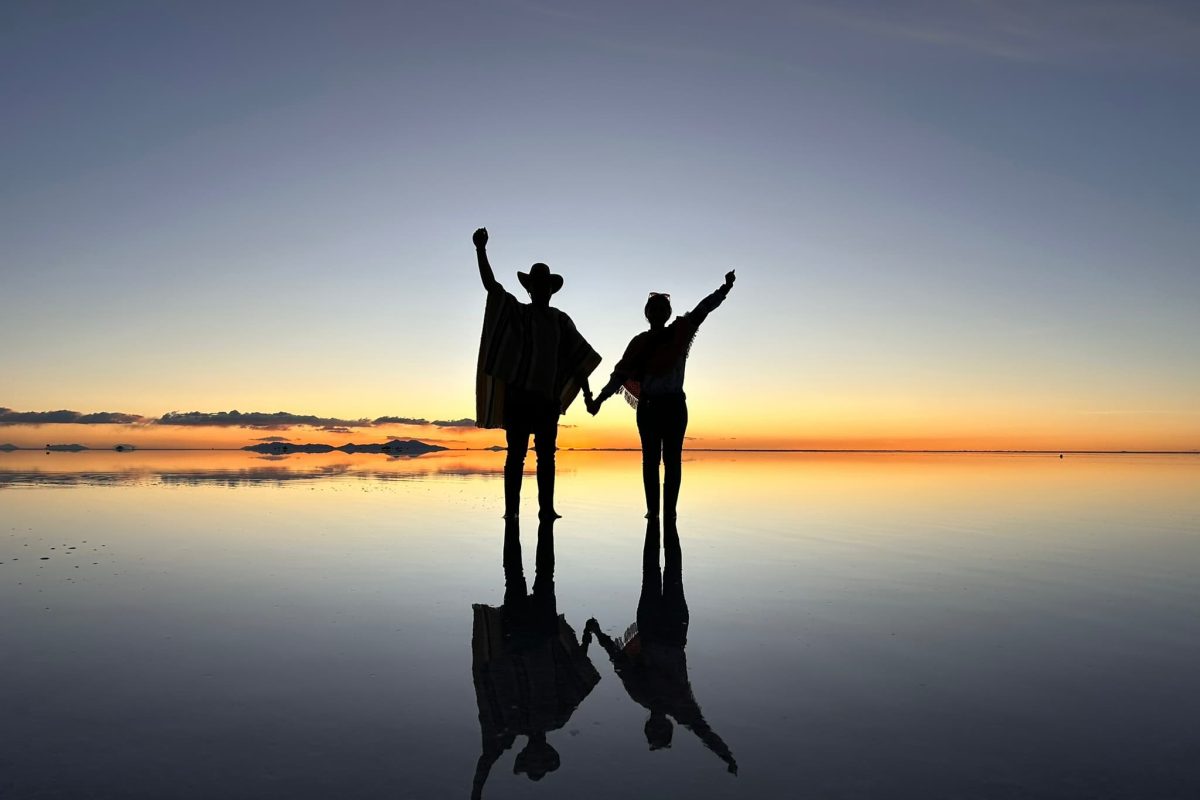 Siluetas de pareja tomadas de la mano con el efecto espejo durante el atardecer en el Salar de Uyuni, Bolivia